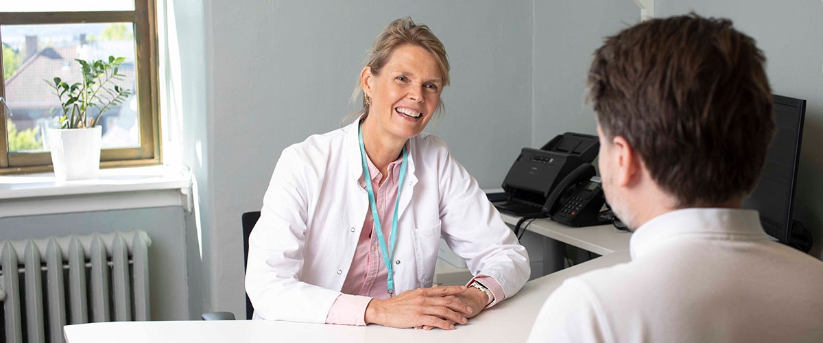 smilende kvinne i legefrakk konsulterer mannlig pasient på legekontor. 